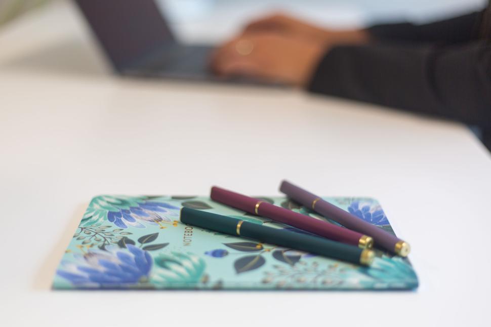Free Image of Three colorful pens on closed floral notebook with person using laptop. 