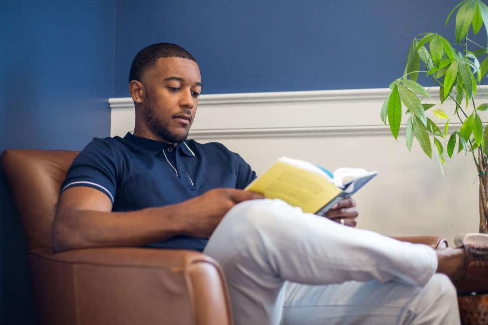 Free Image of Man reading book in brown armchair against blue wall indoors 