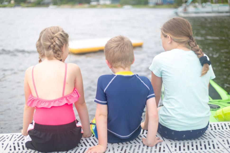 Free Image of Three children sitting by the lakeside with water and dock visible. 
