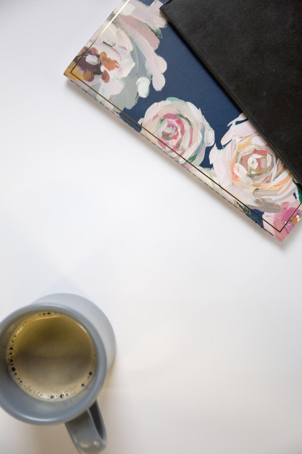 Free Image of Coffee cup next to floral notebook on a white surface, from above. 