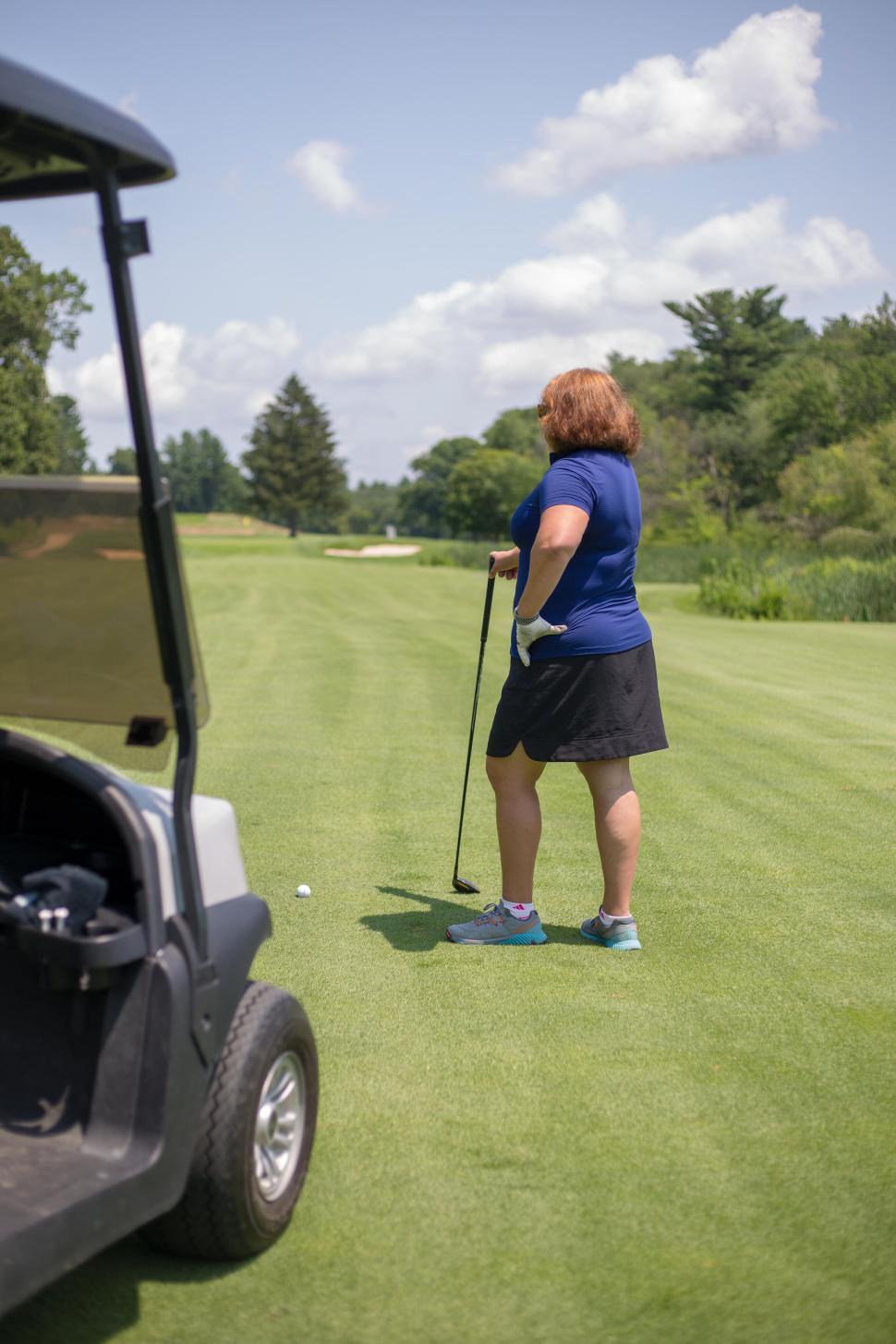Free Image of Woman on golf course, standing with a golf club next to a cart. 