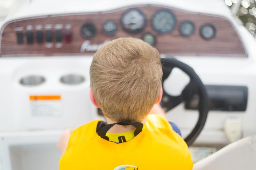 Free Image of Young child steering a boat, back view wearing yellow life jacket 