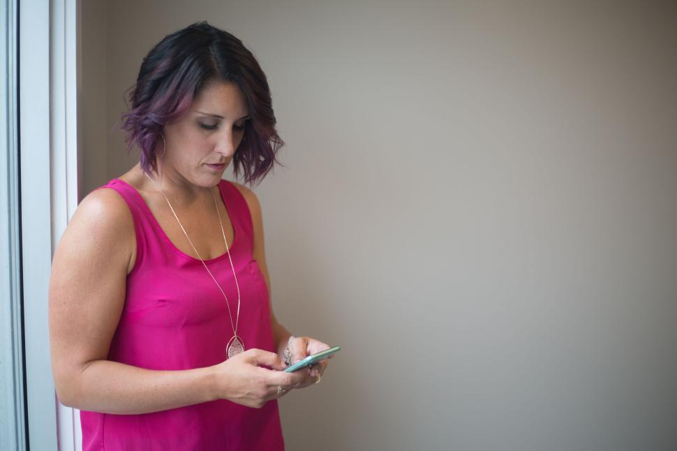 Free Image of Woman in pink top using smartphone by window in modern setting. 