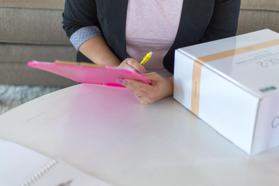 Free Image of Person writing with a pen on a clipboard beside a box on a table. 