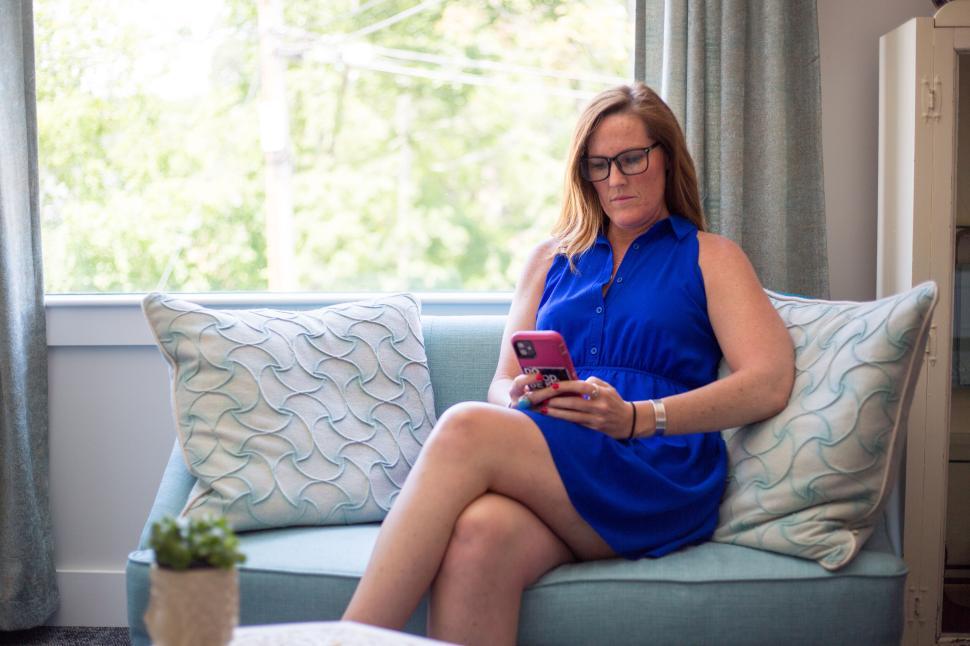 Free Image of Woman in blue dress using phone on sofa near window indoors. 