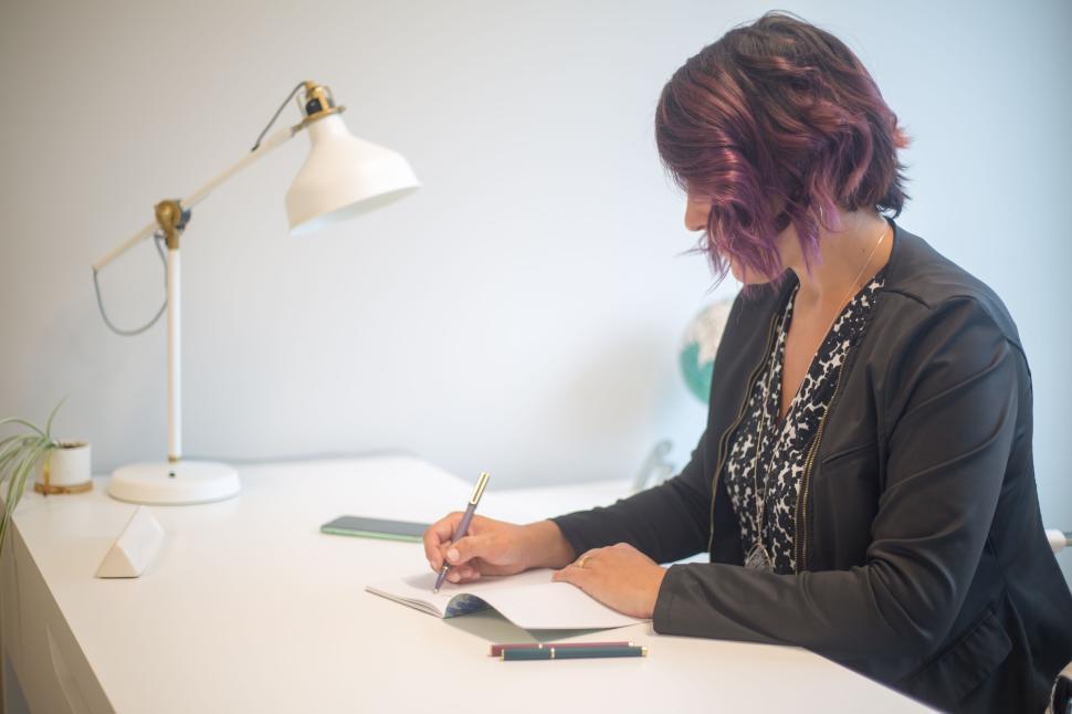 Free Image of Woman writing notes at her desk with modern office lighting 