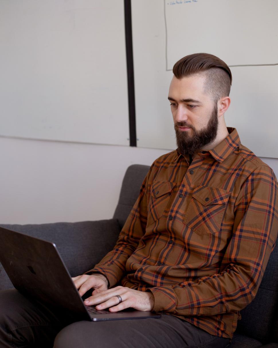 Free Image of Bearded man in plaid shirt using laptop seated on gray sofa. 