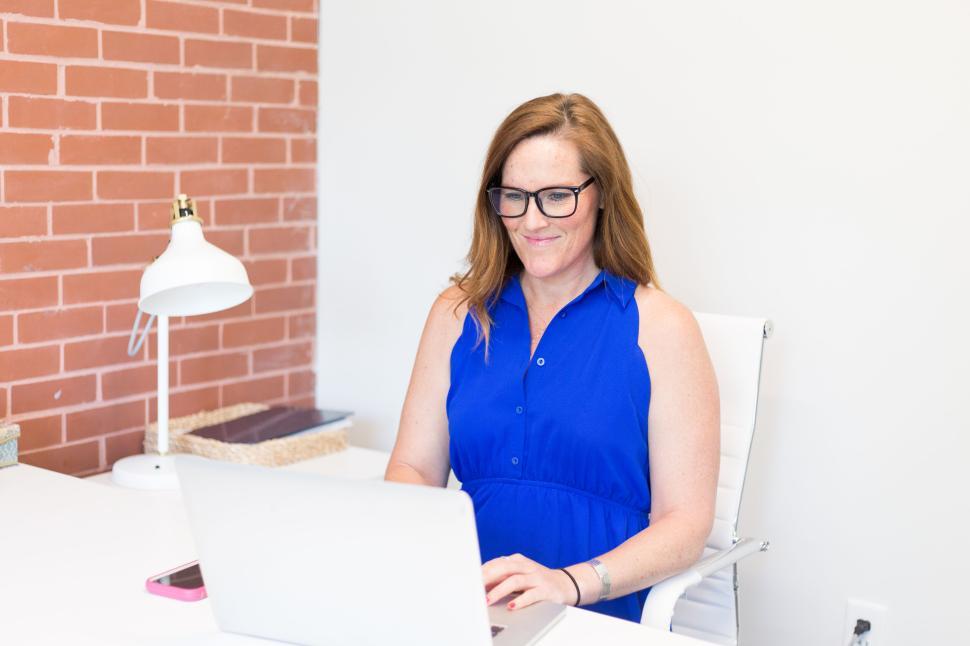 Free Image of Woman in blue dress working on laptop at white desk in office. 