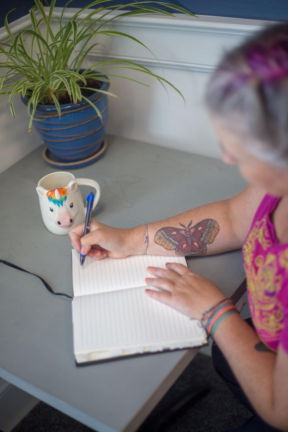 Free Image of Person writing in a notebook beside a plant and a whimsical mug 