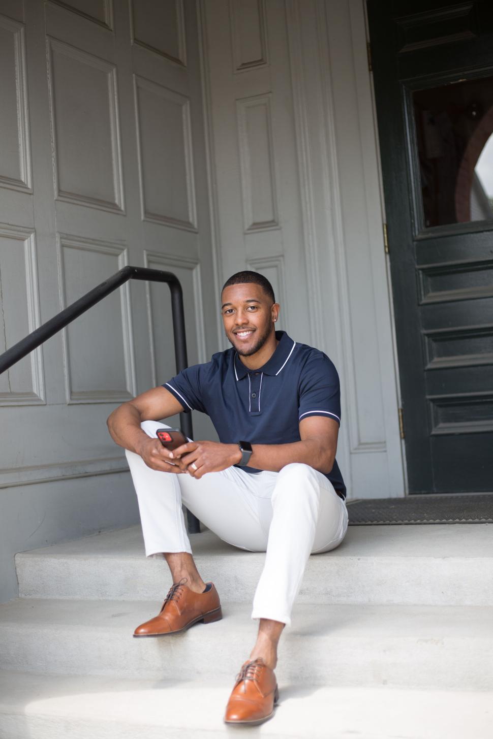 Free Image of Smiling man seated on steps, holding a phone, in a casual outfit. 