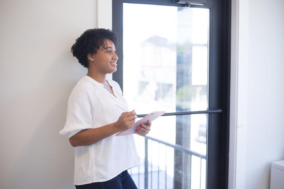 Free Image of Woman standing by a window, holding a tablet and smiling. 