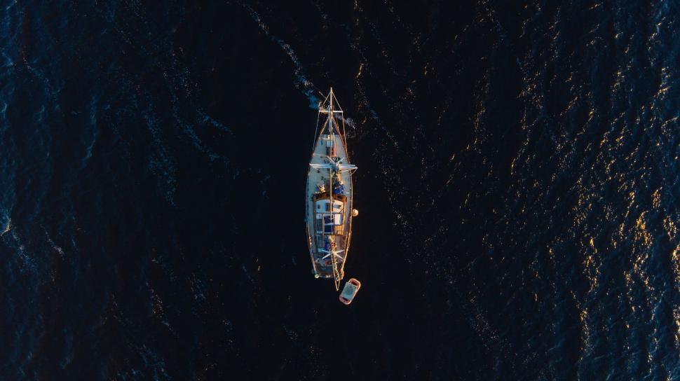 Free Image of Aerial shot of a sailboat in dark blue waters with a small dinghy. 