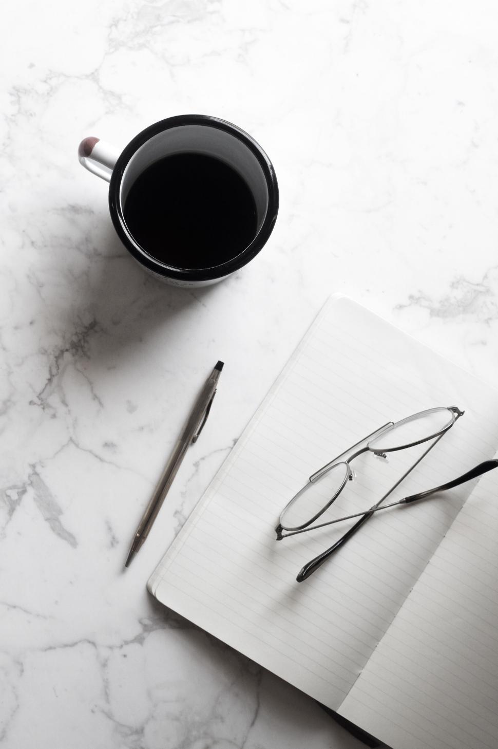 Free Image of Coffee mug, open notebook with pen and glasses on a marble table. 