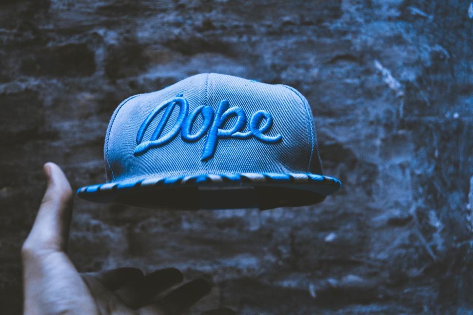 Free Image of Grey cap with  Dope  text written on it held by one hand in dim light. 