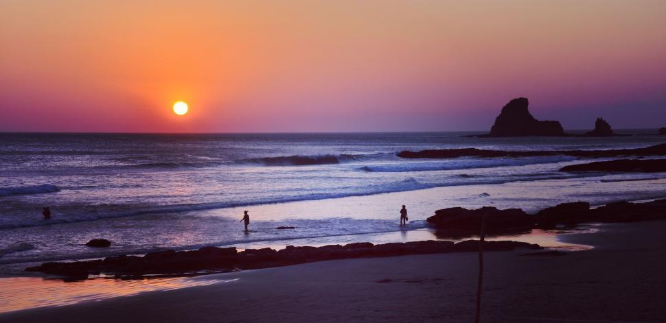 Free Image of Sunset on a tranquil beach with figures wading in the water 