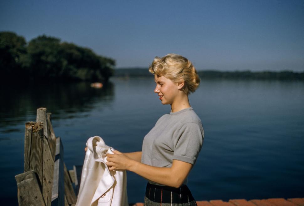 Free Image of Young woman holding white cloth beside lake with clear blue sky. 