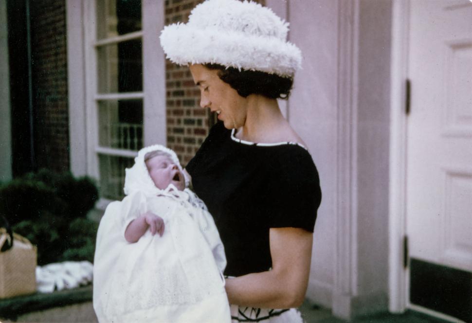 Free Image of Woman in stylish hat holds newborn baby outside residential building. 