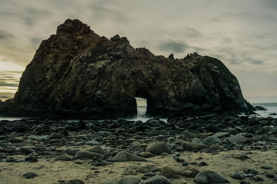 Free Image of Rock formation with hole in the middle on a pebble-filled beach. 