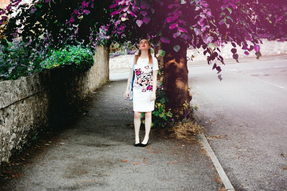 Free Image of Woman standing under tree in white dress on a quiet street day 