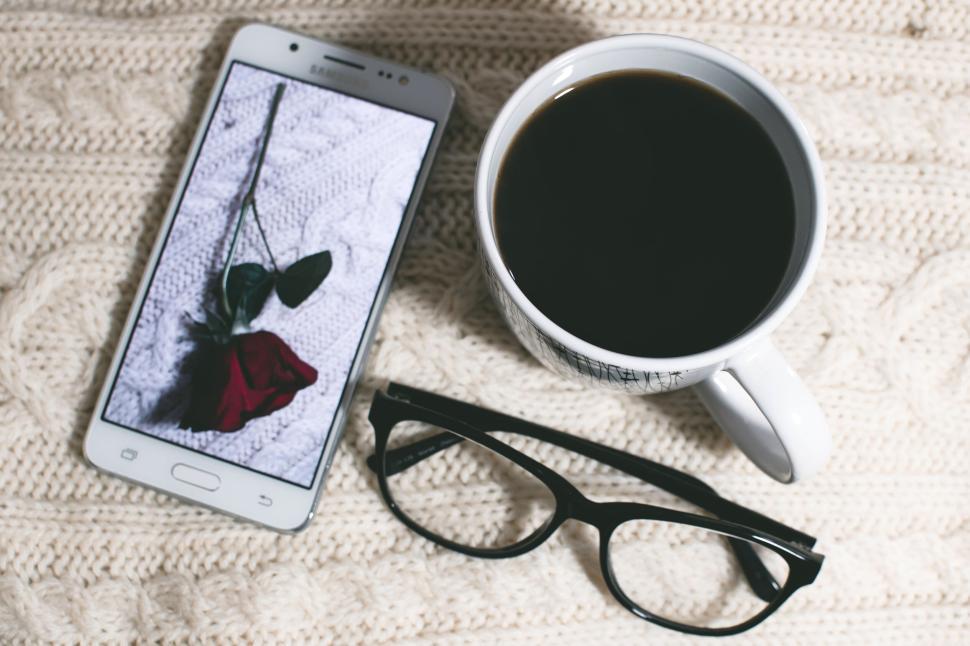 Free Image of Coffee, smartphone displaying a rose, and eyeglasses on knitted fabric. 