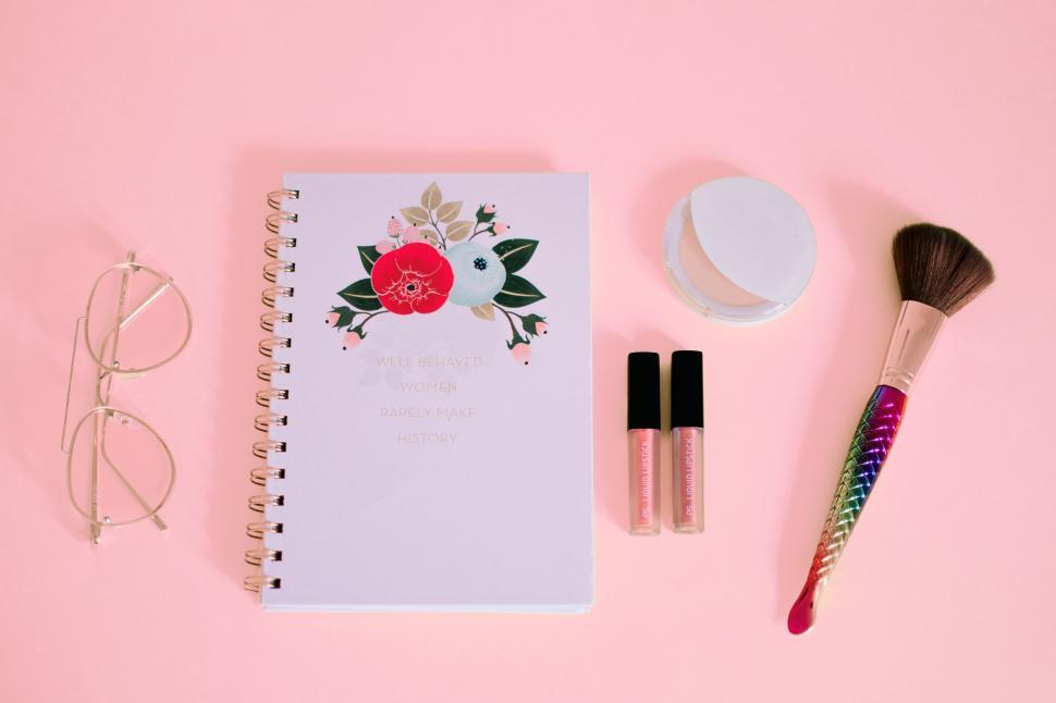 Free Image of Flat lay of notebook, glasses, makeup items on a pink background 