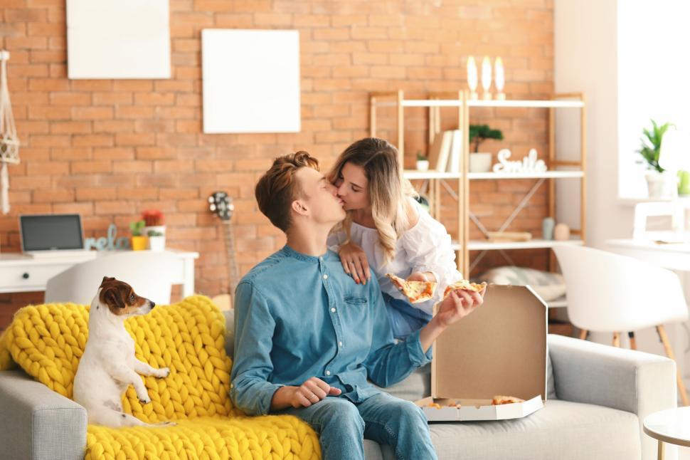 Free Image of Couple sharing a kiss with pizza on a cozy living room couch 