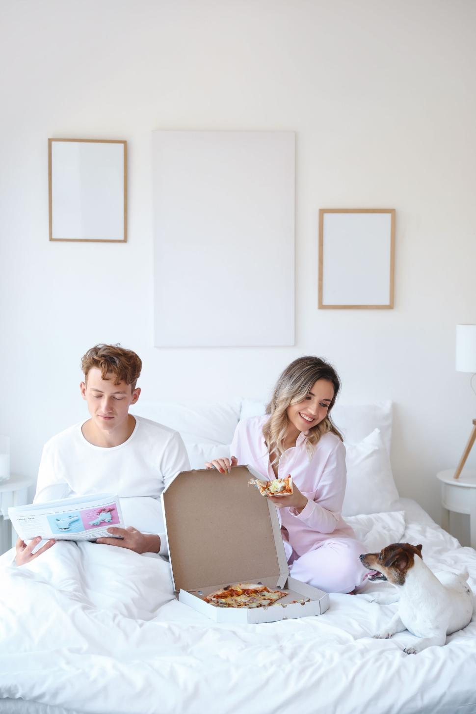 Free Image of Couple enjoying pizza in bed with a curious dog by their side 