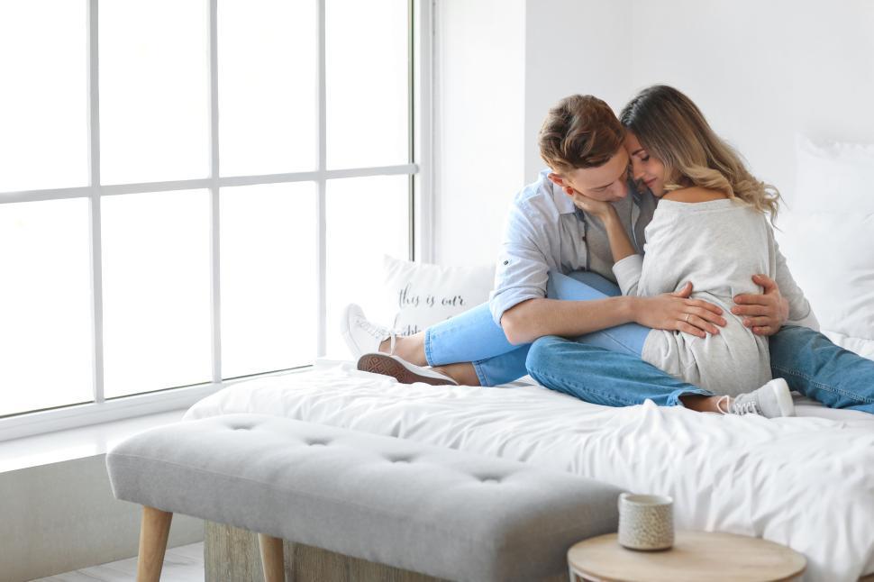 Free Image of Couple sitting on white bed embracing with a window in the background 