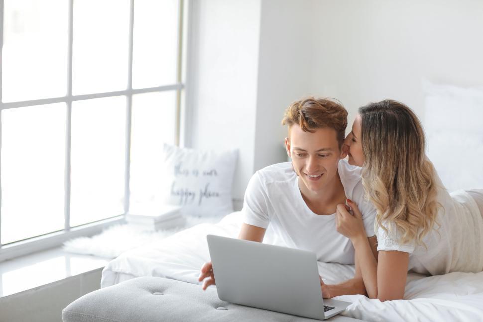 Free Image of Couple with laptop, woman whispering in man s ear, cozy bedroom. 
