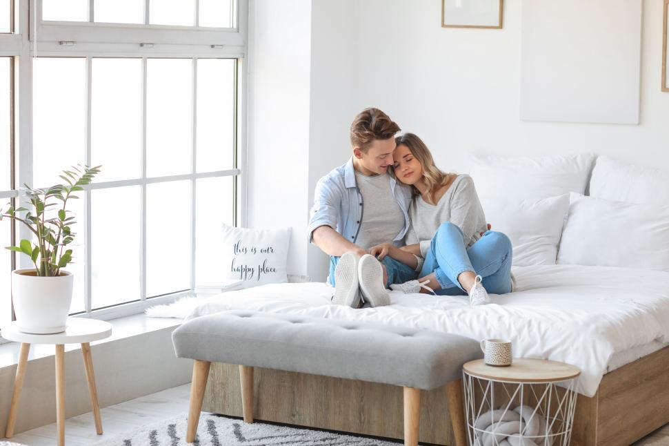 Free Image of Couple sitting close together on a bed in a bright white bedroom 
