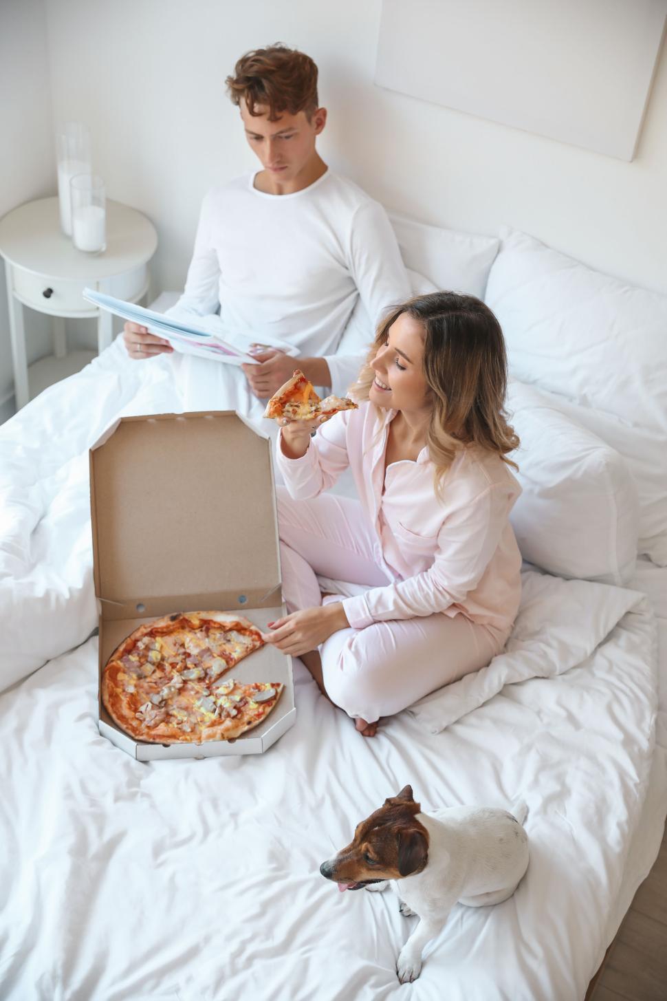 Free Image of Couple in bed enjoying pizza with their dog by their side. 