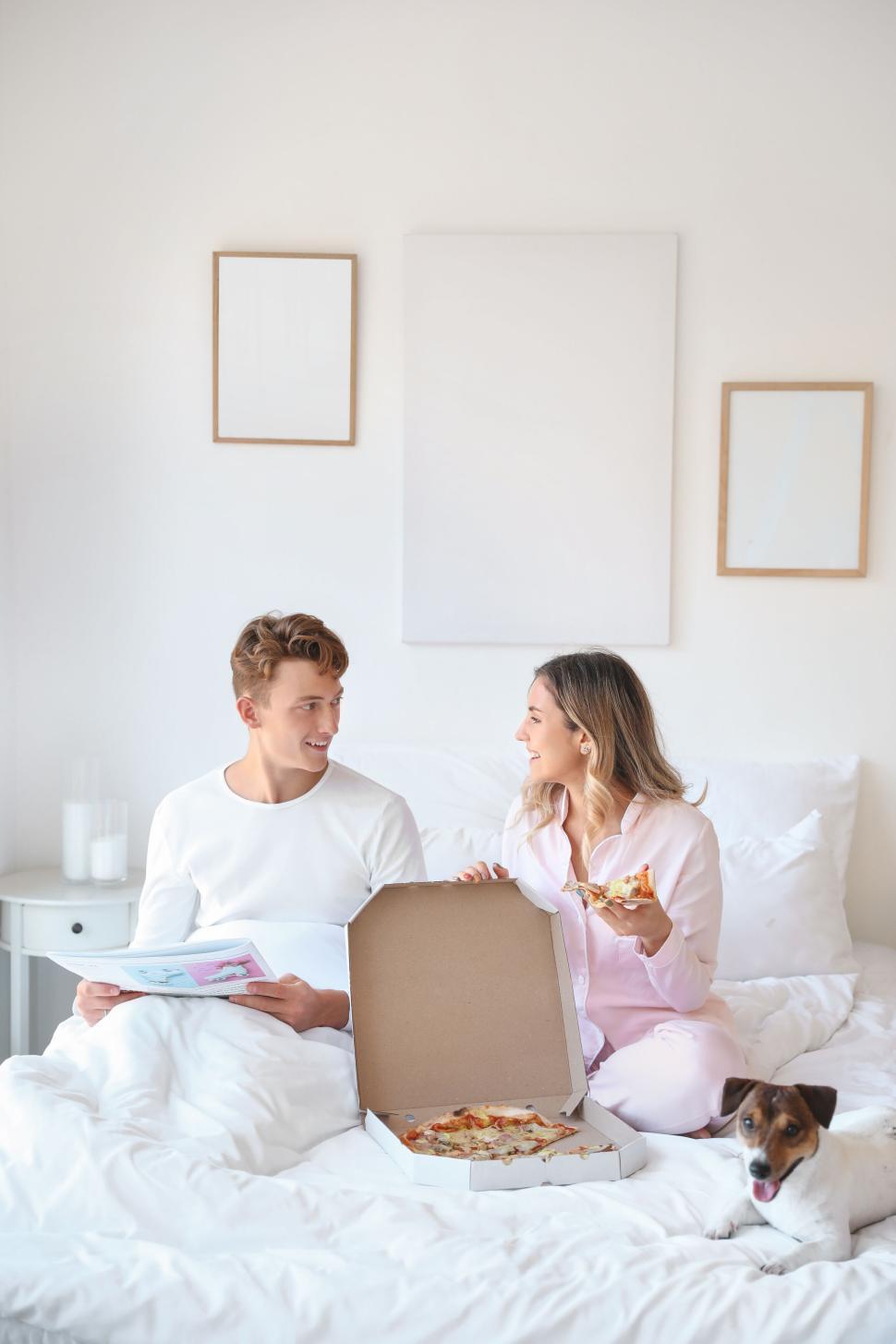 Free Image of Couple enjoying pizza in bed with their dog in a cozy bedroom 