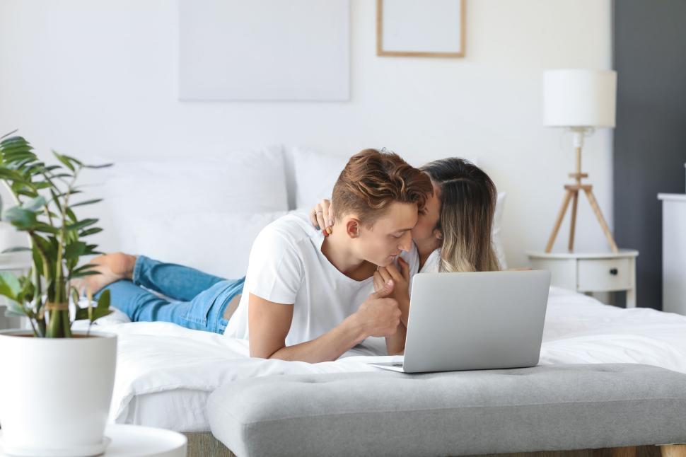 Free Image of Couple lying on bed using laptop with white decor around them. 