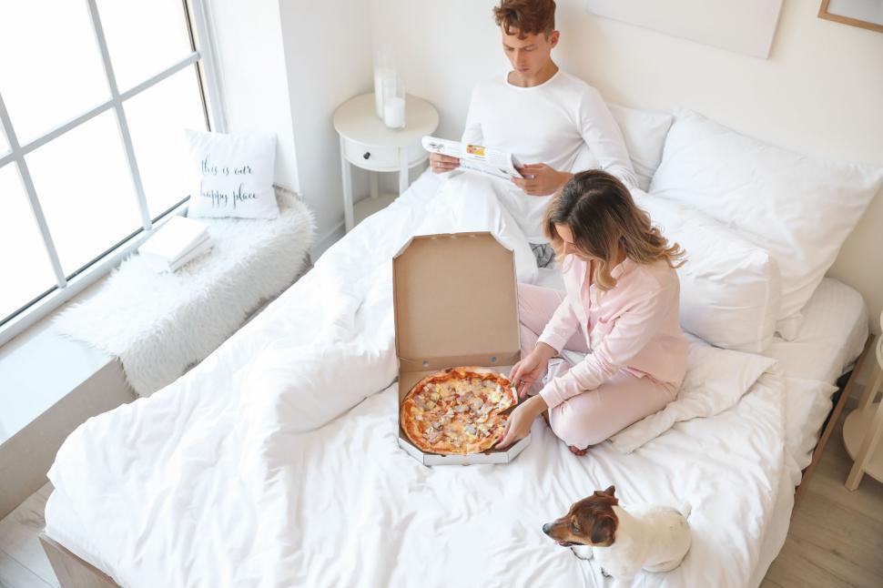 Free Image of Couple with dog enjoying pizza while sitting on the bed together 