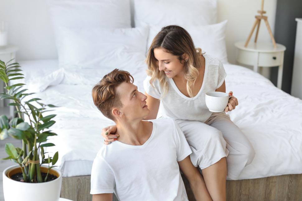Free Image of Couple in white shirts sharing a cup of coffee on the bed. 