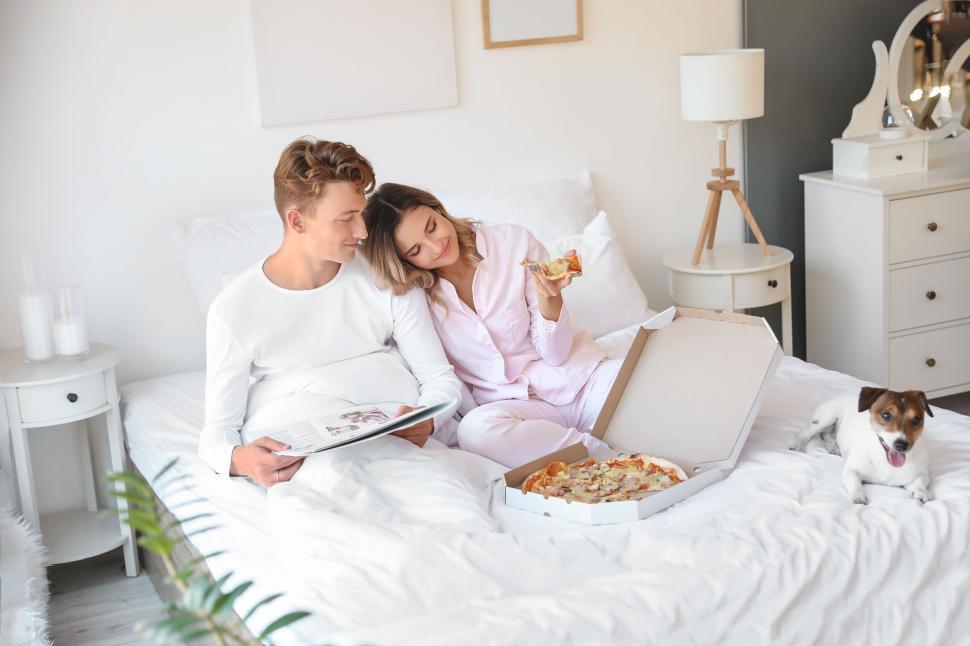 Free Image of Couple eating pizza in bed with dog, sharing a tender fun moment 