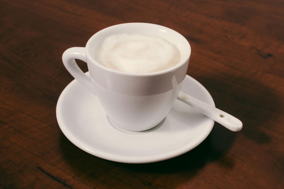 Free Image of White ceramic cup of frothy coffee on a saucer with a spoon on a table. 