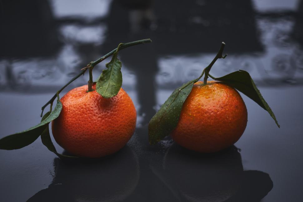 Free Image of Two fresh oranges with green leaves on a reflective dark surface 