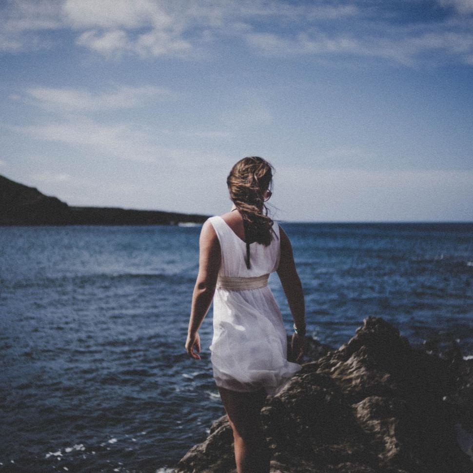 Free Image of Woman in white dress facing the sea on a rocky shoreline 