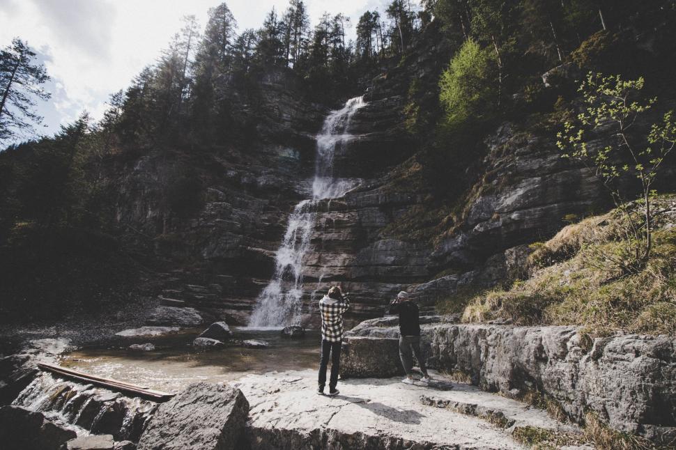 Free Image of Two people standing on rocks photographing a cascading waterfall in forest. 