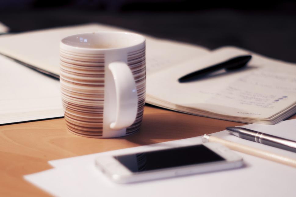 Free Image of A white smartphone, pen, notebooks, and a coffee mug on a desk. 