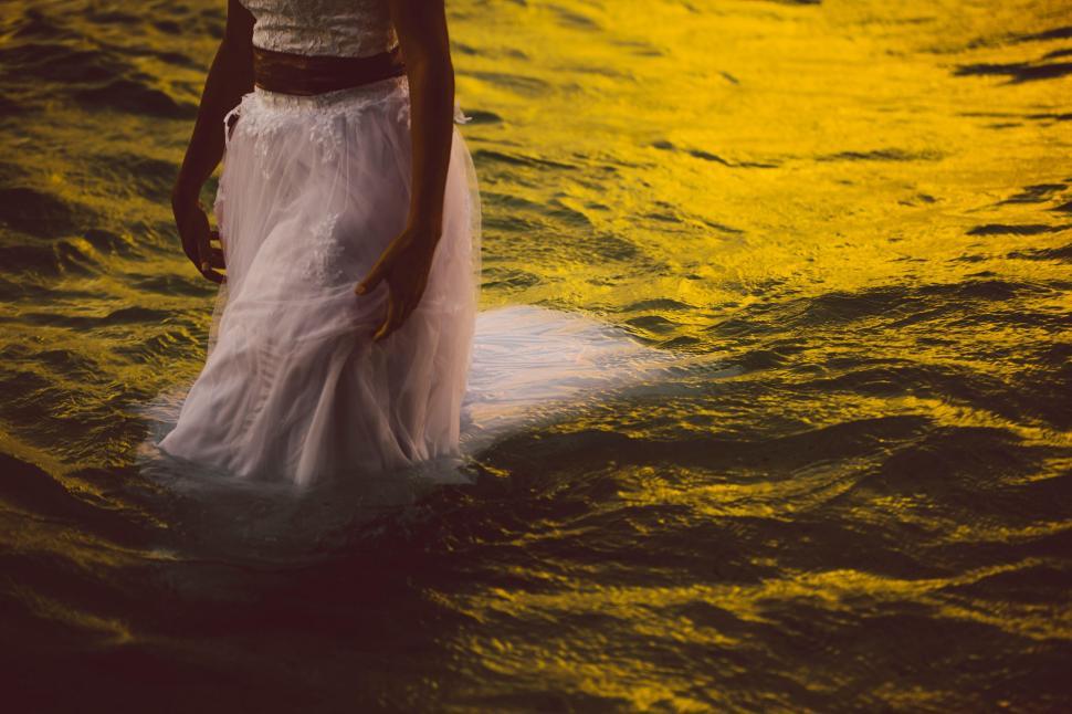 Free Image of Bride in white dress standing in water at sunset with warm glow. 