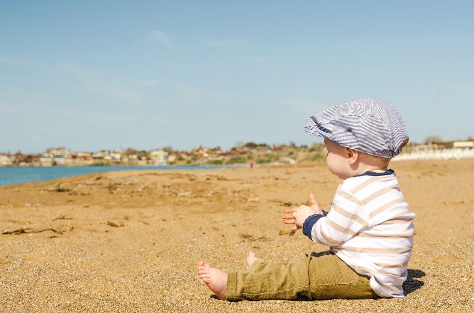 Free Image of Toddler dressed in stripes and cap sits on sandy beach facing ocean 