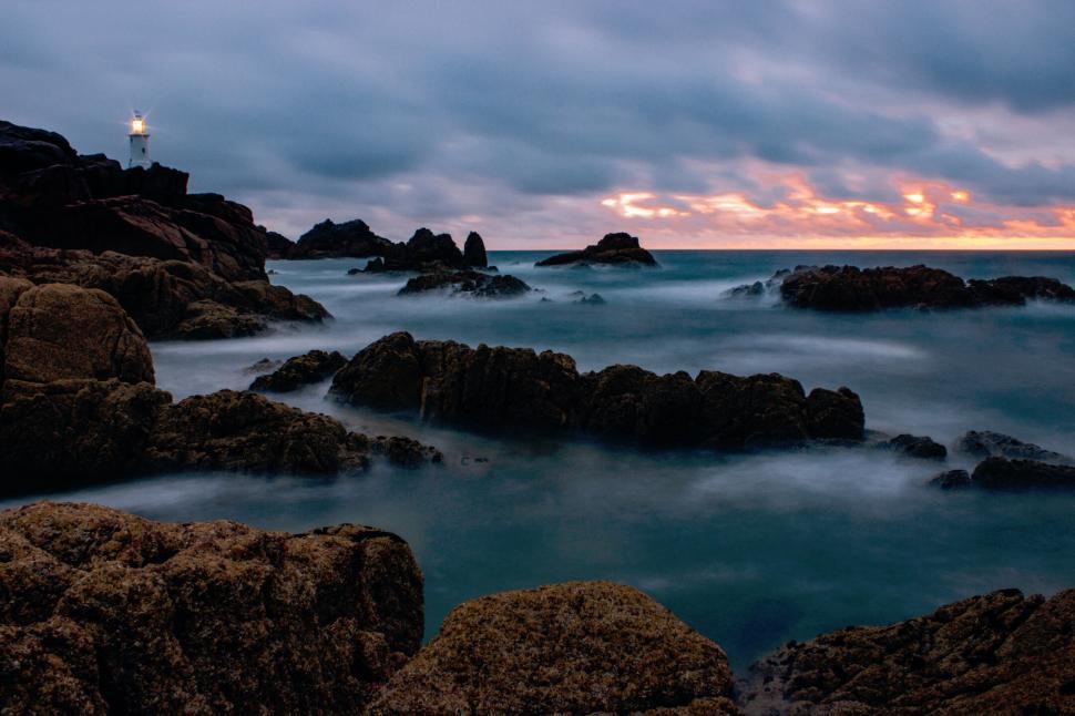 Free Image of Rocky shoreline with lighthouse during colorful sunset over ocean 