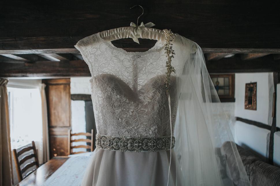 Free Image of White lace bridal gown hanging in rustic, wooden-beamed room. 