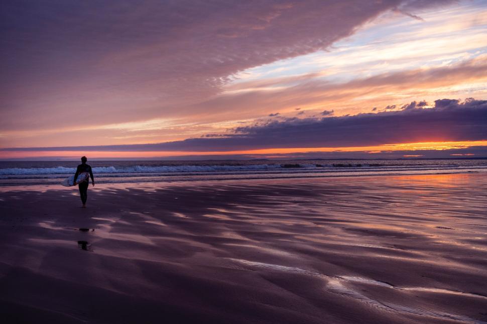 Free Image of Person walks on beach at sunset with surfboard in serene moment 