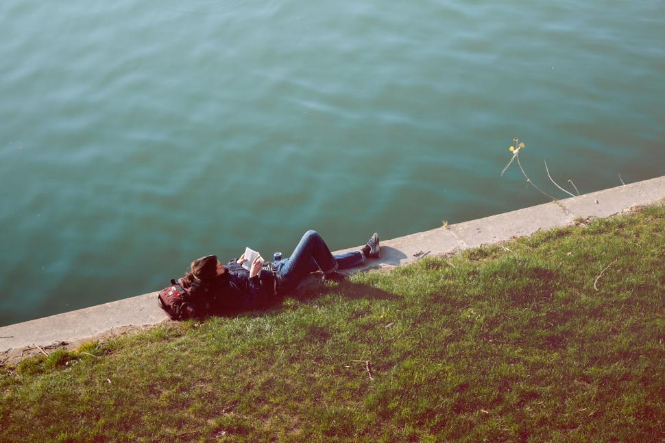 Free Image of Person reading by the water on a sunny day with hat and backpack 