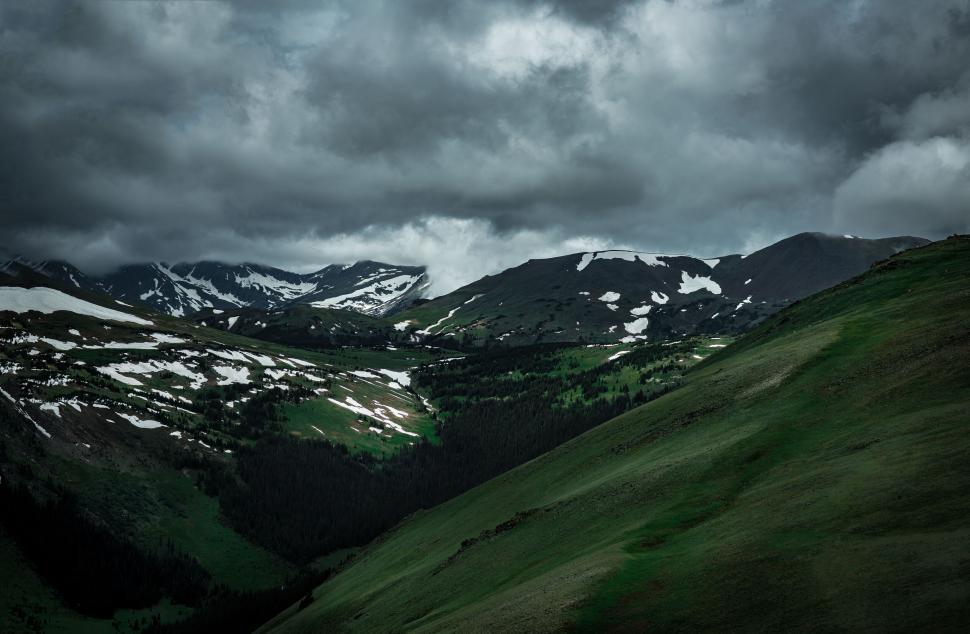 Free Image of Lush mountain valley with snow patches under dark clouds. 