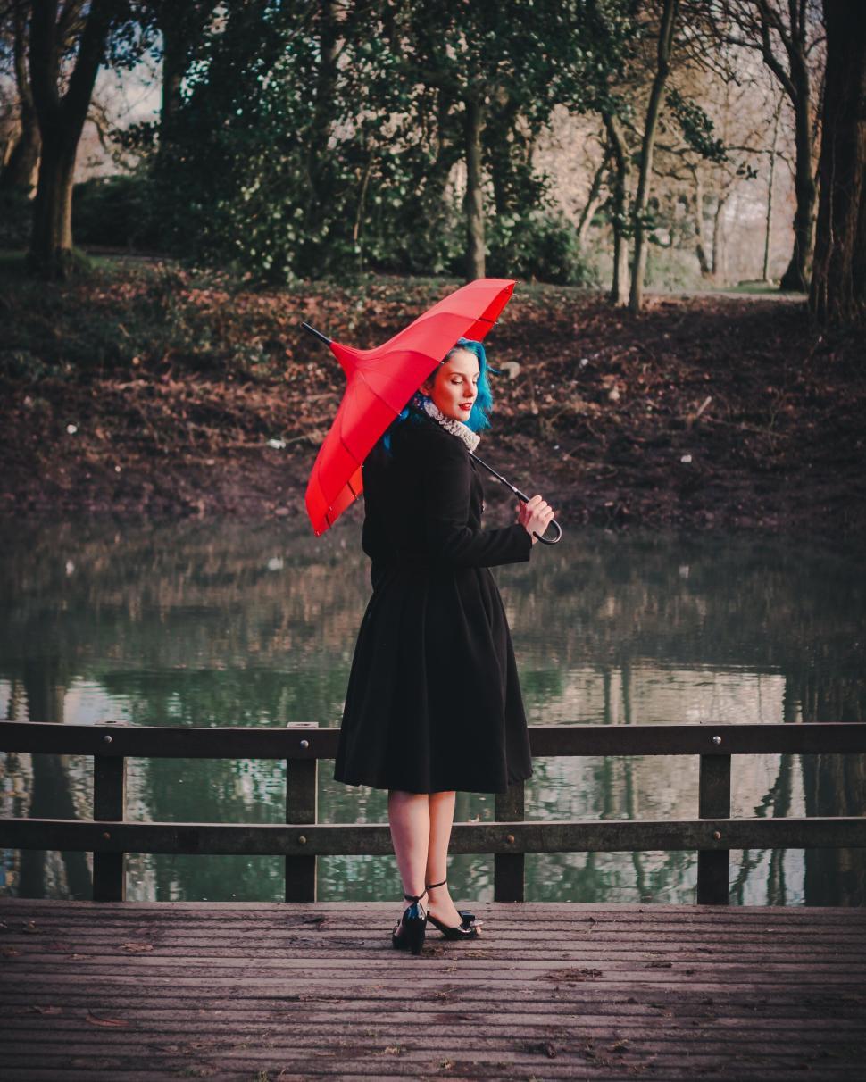 Free Image of Woman with blue hair holds red umbrella near a lake. 