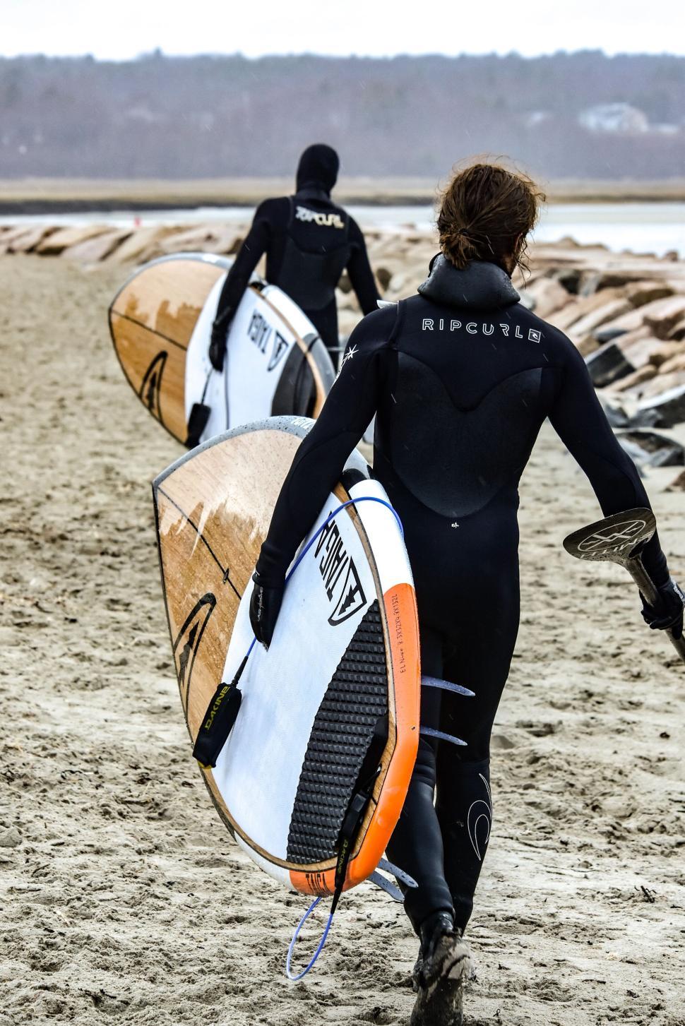 Free Image of Surfers in wetsuits carrying their boards on the beach. 